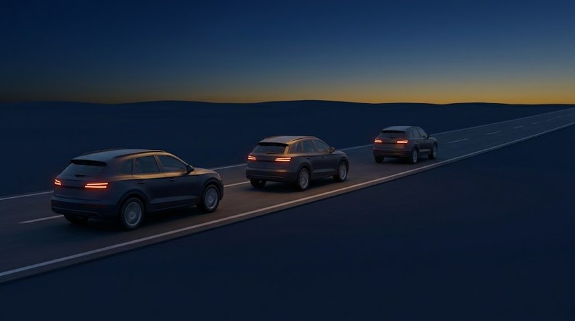 Three cars driving in convoy on an empty highway at dusk, tail lights lit.